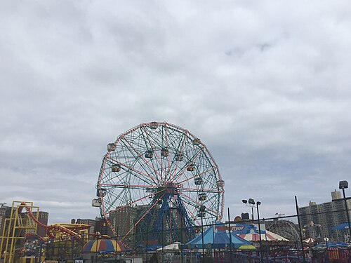 The Wonder Wheel close and centered with its name sign and passenger cars, ride structures and carousel tents of Deno's park below, Cyclone visible at right.