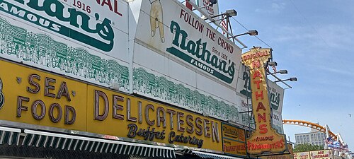 The Nathan's Famous corner on Surf Avenue, neon signage wall and coaster track behind.