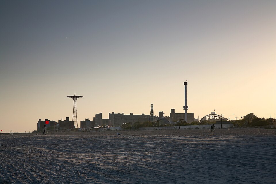 The beach at low light just before sunset, rippled sand in the foreground and the Parachute Jump, Astrotower and amusement skyline silhouetted along the shore, the Warriors-at-dawn mood.
