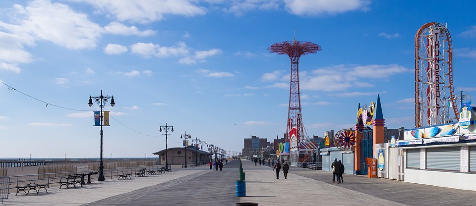 The Riegelmann Boardwalk itself, herringbone boards filling the foreground, ornate lampposts, benches and rail on the beach side, Parachute Jump and the Thunderbolt sign ahead, beach at left.