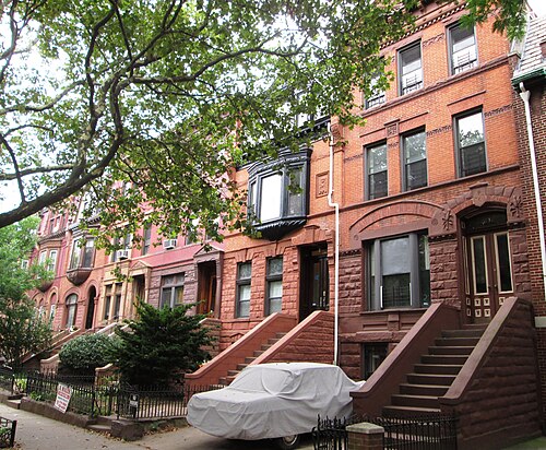 Brownstone houses at 1232 to 1242 Dean Street under a plane tree, carved arches over the doors, a black bay window on one front and high stoops on all of them.
