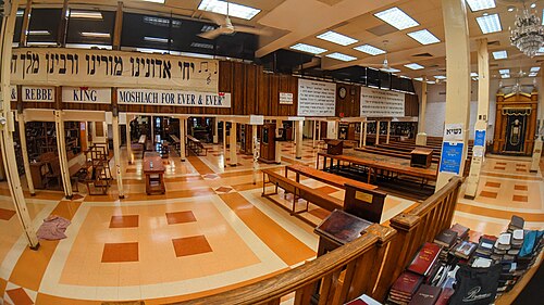 The main hall inside the Lubavitch headquarters at 770 Eastern Parkway, an orange and cream tile floor, plain wooden benches and reading stands, and a printed banner strung along the balcony.