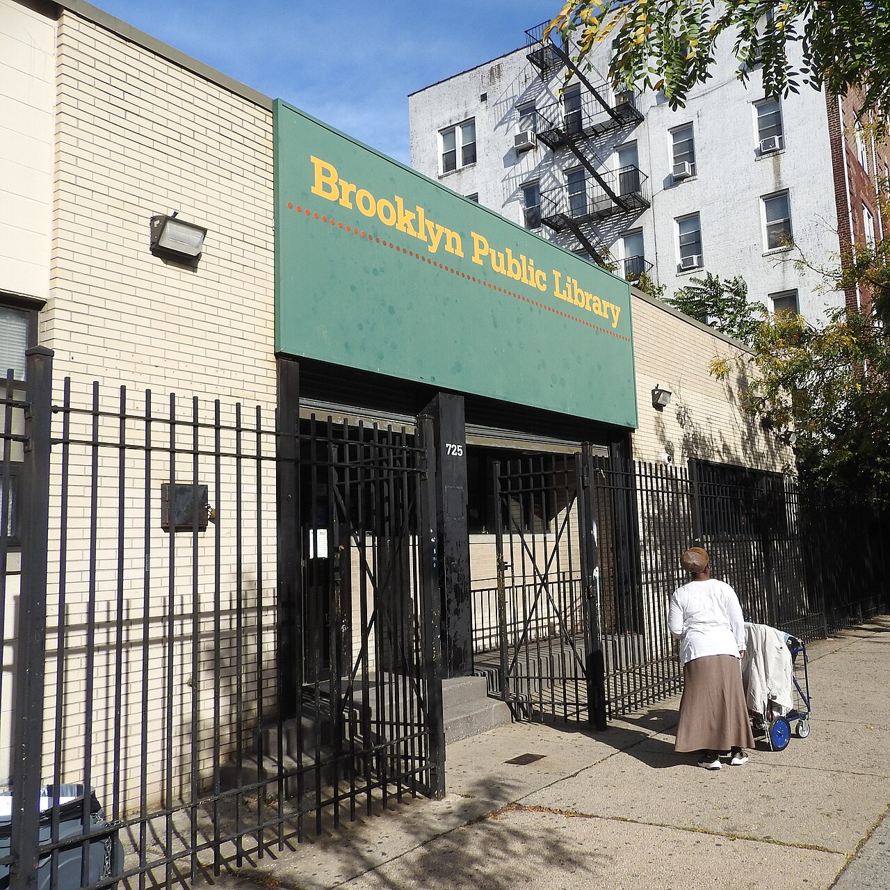 The Brooklyn Public Library branch at 725 St. Marks Avenue, a low cream brick front behind iron gates, with a whitewashed apartment building and its fire escape next door.