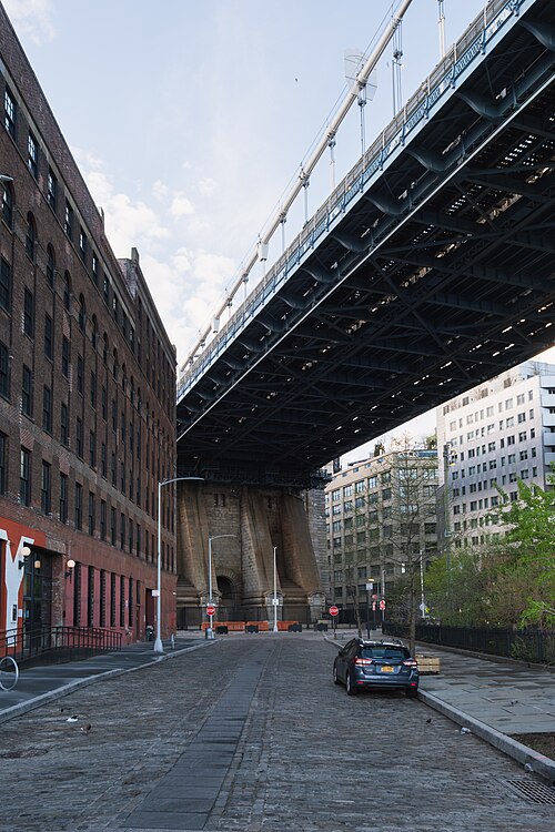 A Belgian block side street under the bridge deck, warehouse left, the granite anchorage closing the view.