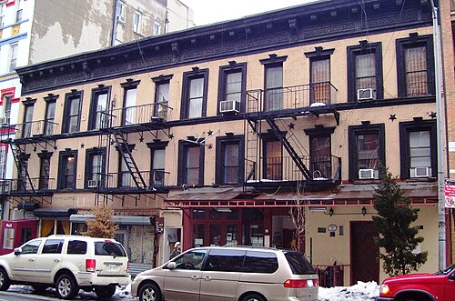 A row of tenements on Avenue B, fire escapes on every front, iron star anchors set into the brick and shops at street level.