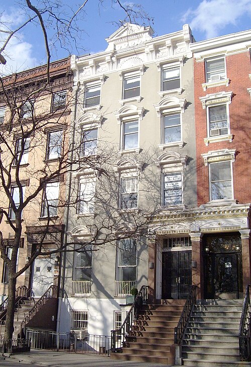 A rowhouse on East 10th Street opposite Tompkins Square Park, pale painted front with a high stoop and a carved doorway, brick houses either side.