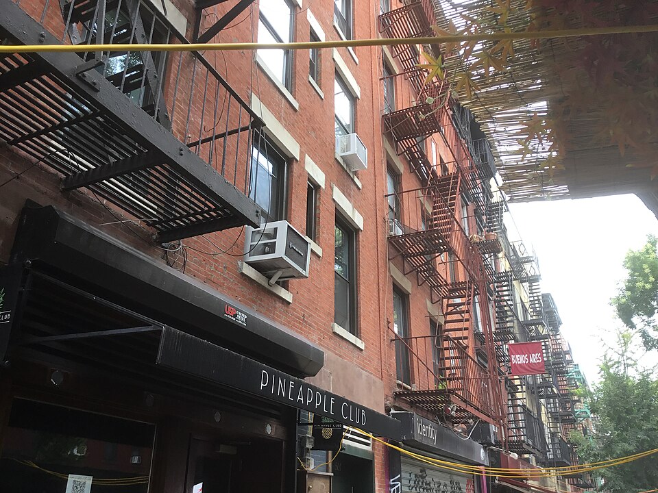 East 6th Street between Avenue A and Avenue B, red brick tenements with fire escapes running up the fronts and small storefronts at the sidewalk.
