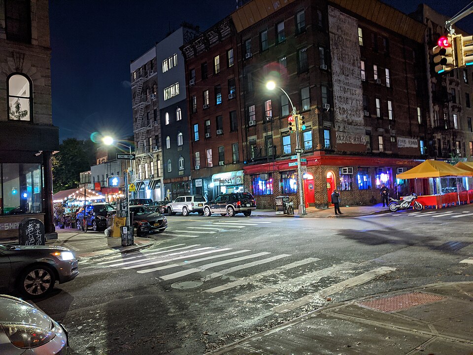 The corner of East 7th Street and Avenue B at night, tenements round the intersection and the bar lit up in red on the corner.