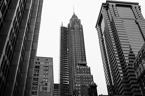 A low angle up a narrow block at an Art Deco tower with a stepped crown and a spire, scaffolding on the building beside it, in black and white.