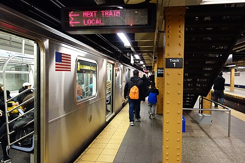 The platform at Flushing Main Street, the eastern end of the 7 line, with riveted yellow columns, a train standing on track 1 and passengers walking toward the stairs.