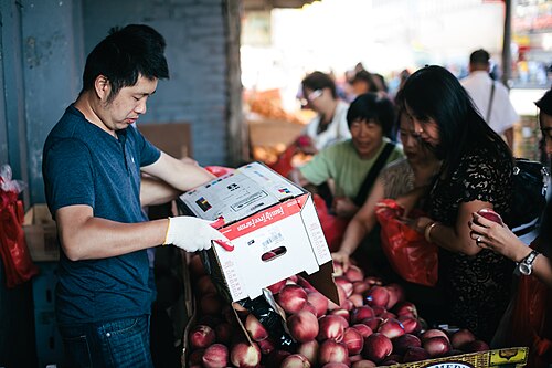 A vendor tipping a box of nectarines onto a heaped table under the Long Island Rail Road bridge, with shoppers picking through the fruit and daylight at the end of the underpass.