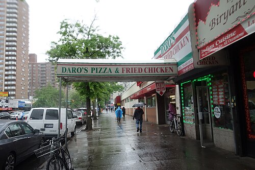 Kissena Boulevard at Elder Avenue in the rain, a long green and red awning over the sidewalk for a pizza and fried chicken shop, a halal biryani place next door and brick apartment towers behind.