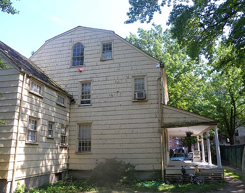 Kingsland Homestead, a clapboard house with a gambrel roof, small pane sash windows and a white columned porch, standing in Weeping Beech Park with brick apartment buildings visible past it.