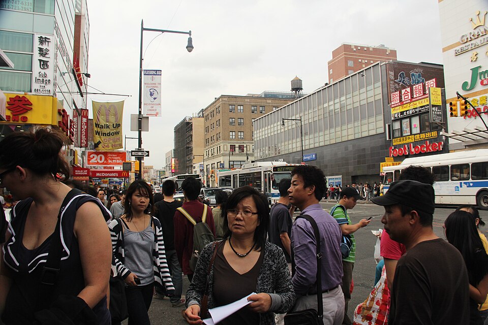 The sidewalk at Main Street and Roosevelt Avenue with a crowd waiting to cross, hanging Chinese shop signs stacked up the building fronts, a twenty four hour walk up window and a wooden water tank on a roof behind.