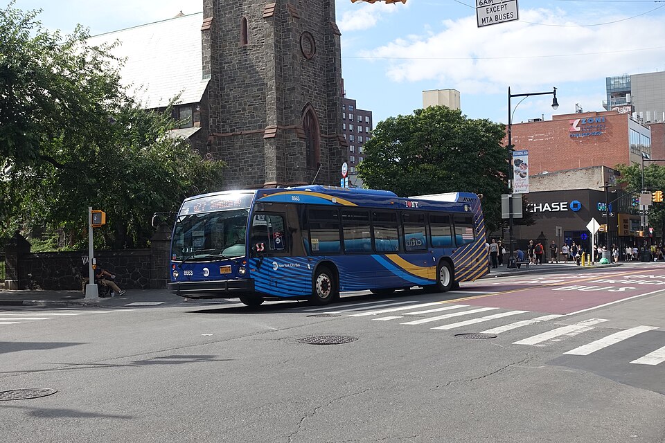 Main Street at 39th Avenue, with a stone church tower on the corner, a bus turning across the red bus lane and shopfronts and crowds along the far side.