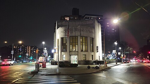 A curved white Art Deco bank building lit up at night on a Forest Hills corner, apartment towers behind it and cars passing in both directions.
