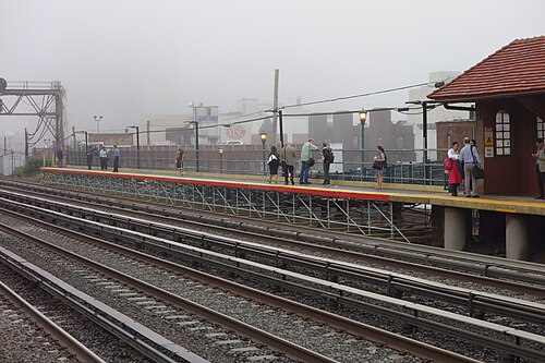 The Long Island Rail Road platform at Forest Hills in heavy fog, commuters waiting near the station house's red tile roof.