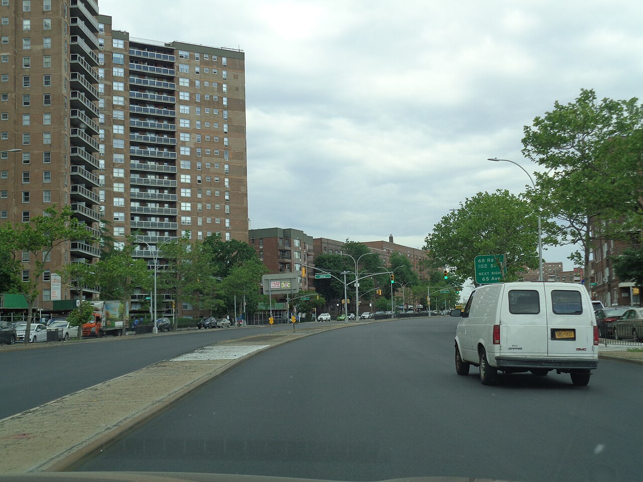 Queens Boulevard in Forest Hills, a tall brick apartment tower on the left, more apartment buildings down the block, and a green highway sign overhead for the next side streets.