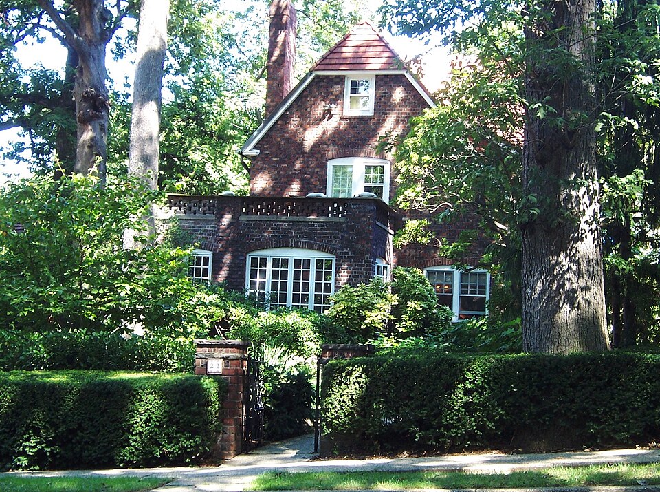 A brick and stucco Tudor house in Forest Hills Gardens, a steep gabled roof and arched ground floor windows behind a clipped hedge and brick gatepost.