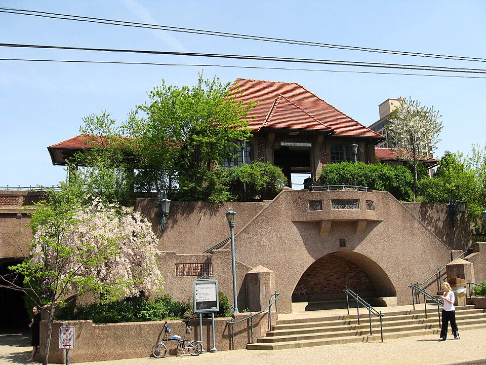 The archway into Forest Hills Gardens beside the Forest Hills train station, a stucco building with a red tile roof above a stone underpass, steps and streetlamps at plaza level.