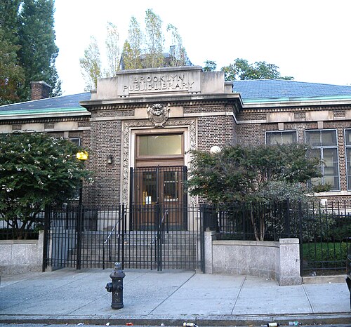 The Walt Whitman branch of the Brooklyn Public Library on St Edwards Street, a low brick and limestone Carnegie library with a green tiled roof behind an iron fence.