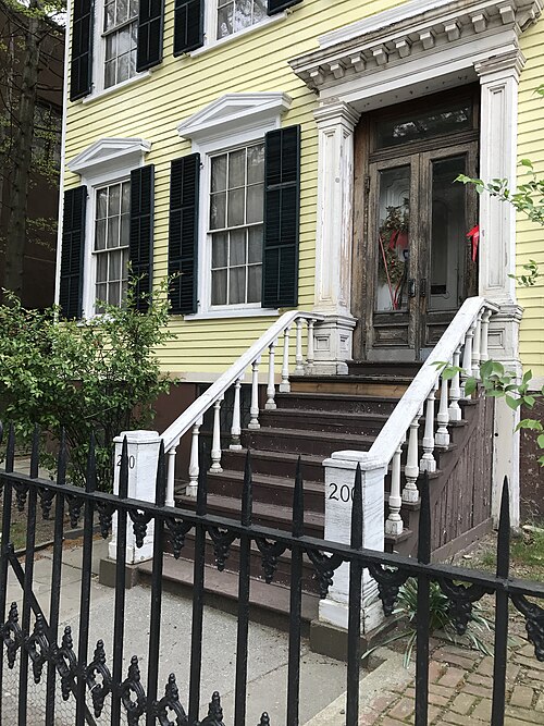 A yellow clapboard house with black shutters and a wooden stoop, the door surround painted white and the double doors left as bare wood.