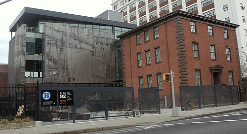Building 92 on Flushing Avenue at the Brooklyn Navy Yard, a brick house with a grey glass and perforated metal addition against its side, behind a black fence.