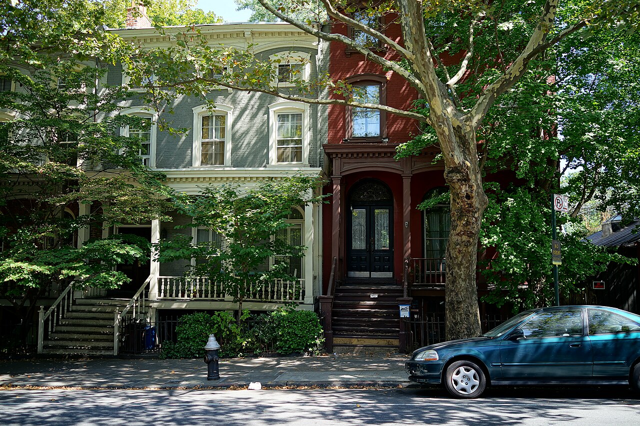 A block on Vanderbilt Avenue seen from across the road, two Italianate rowhouses with high stoops and a plane tree over the sidewalk in deep summer shade.