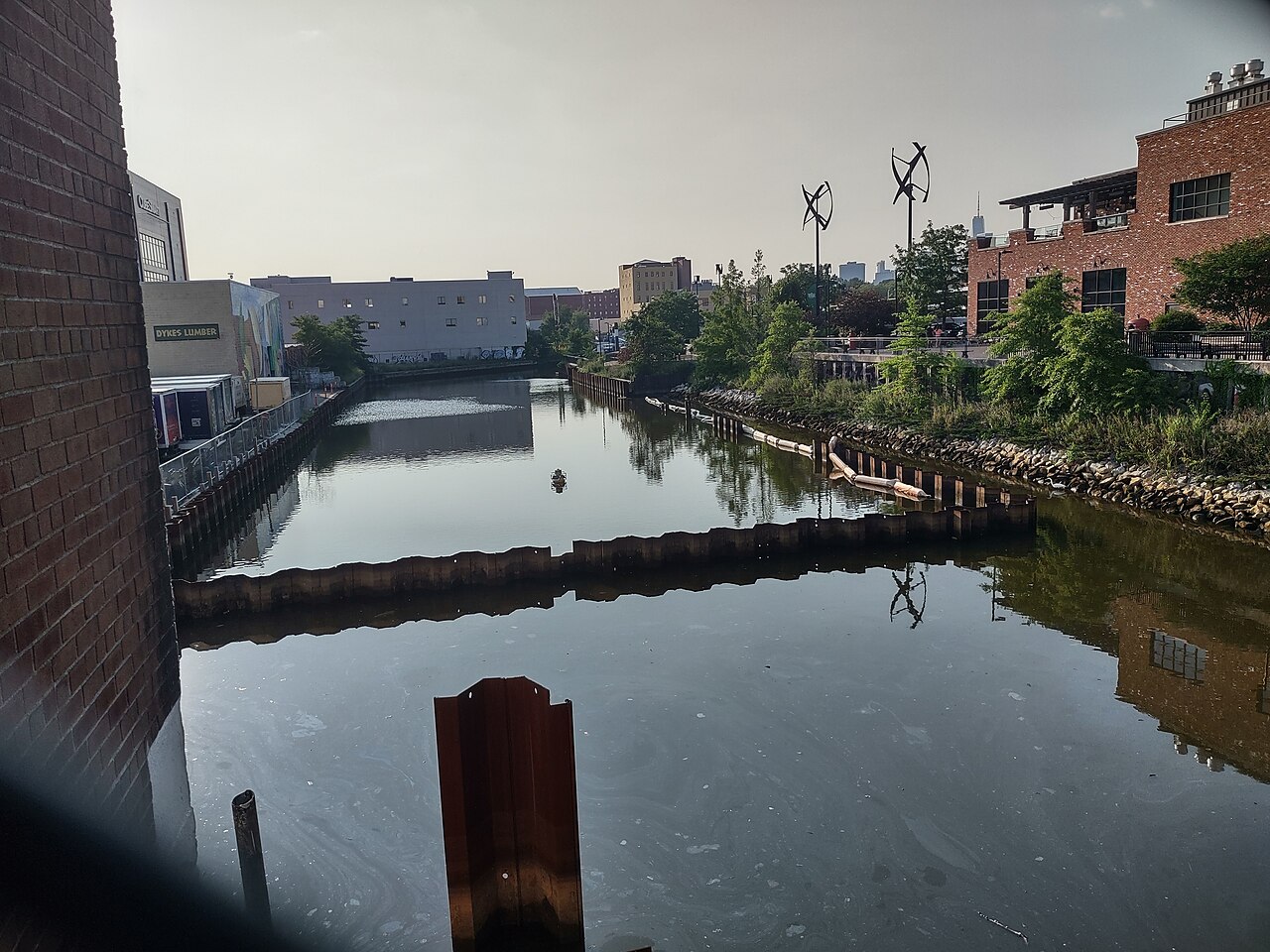 The canal at the Fourth Street Basin, rusted sheet piling along both banks, a lumber yard on the left and two small wind turbines on the far side.