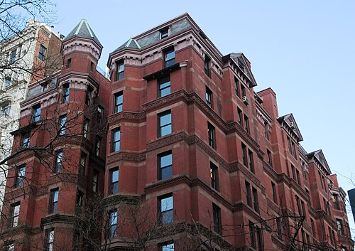 The upper storeys of an apartment building in dark red brick, corner turrets with conical caps, bay windows and a run of dormers along the cornice.