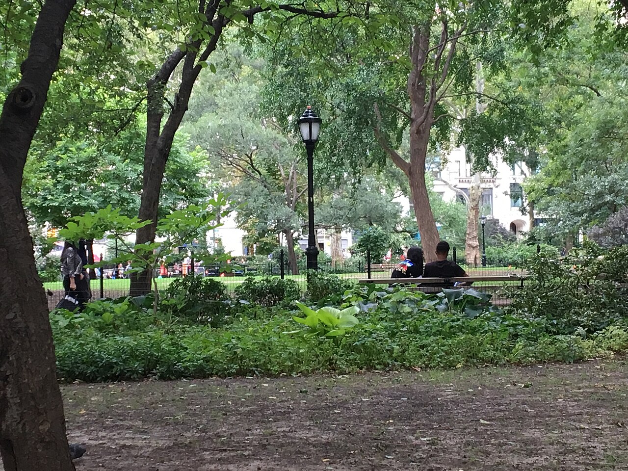 Inside the fence, a bench under tree cover, a black iron lamppost on the gravel path and a brick and stone facade visible through the leaves across the square.