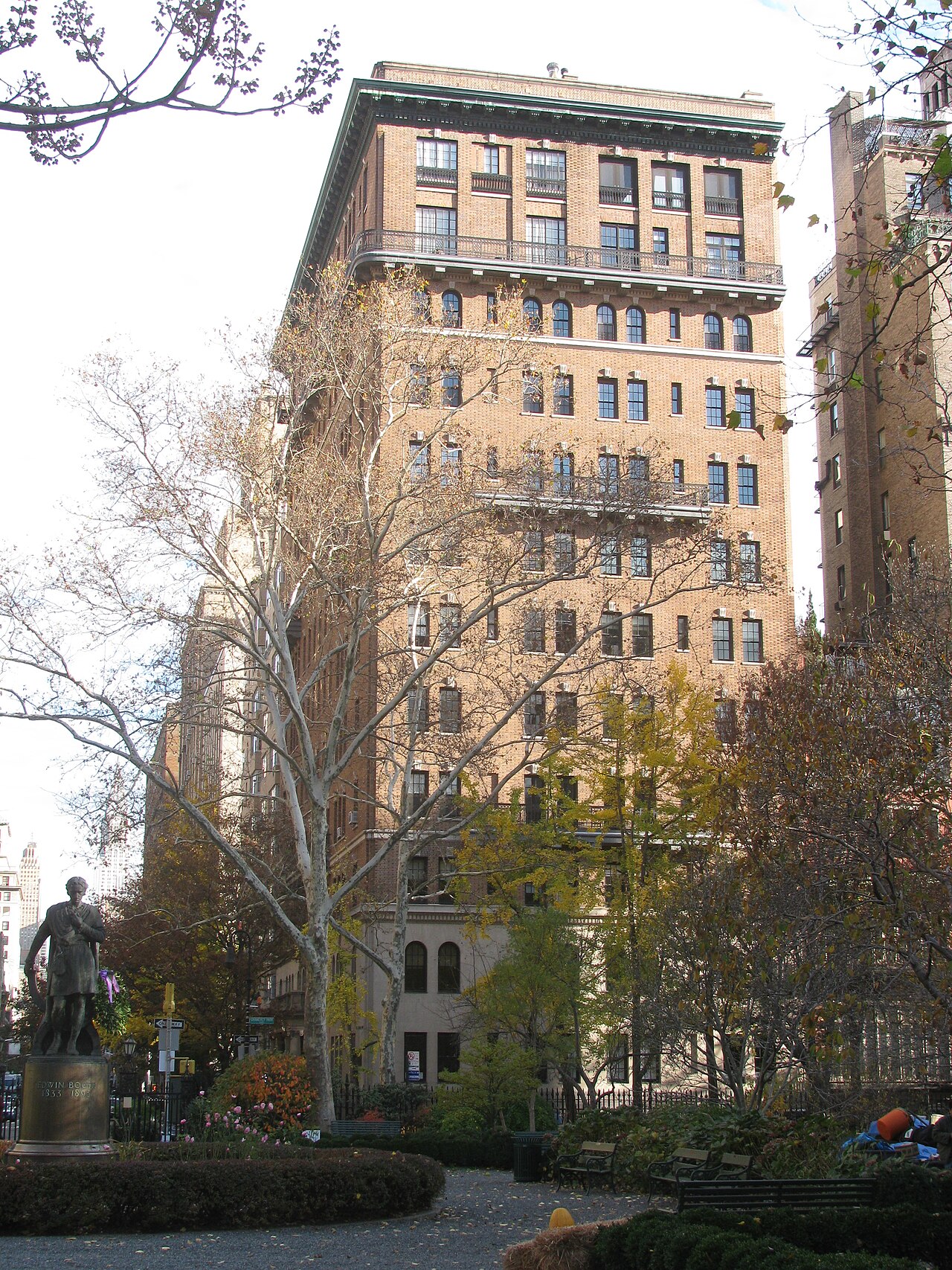 The bronze statue of Edwin Booth among bare trees, park benches along the path and a tall brick apartment building rising behind the fence.