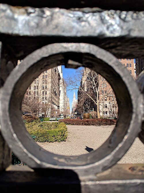 Looking through a round opening in the fence's cast ironwork at a bronze statue and hedges inside the square, apartment buildings and a distant tower beyond.