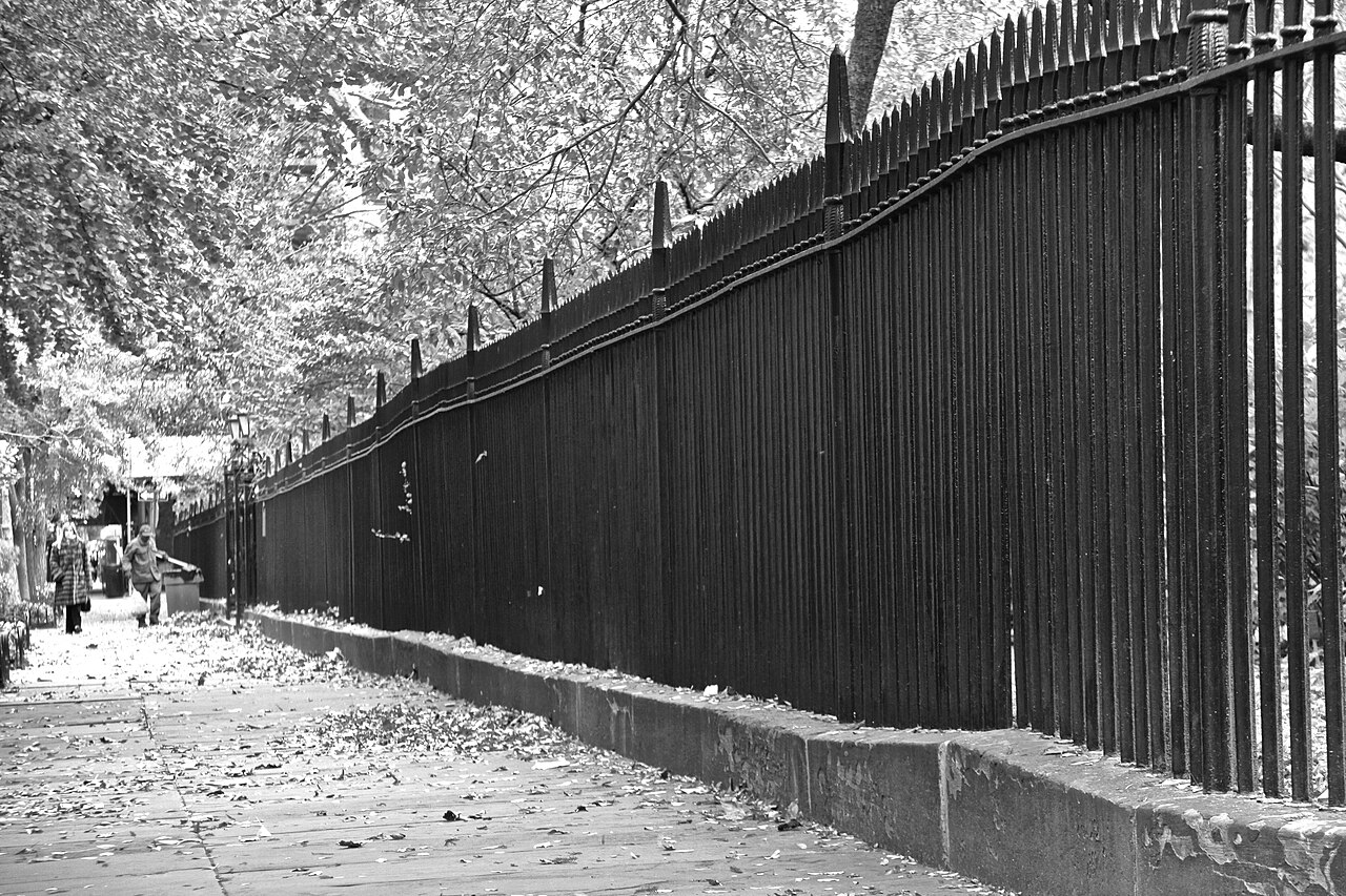 The cast iron fence running along the sidewalk under bare plane trees, its finials repeating post to post the length of the block, autumn leaves banked against the curb and two people walking past.