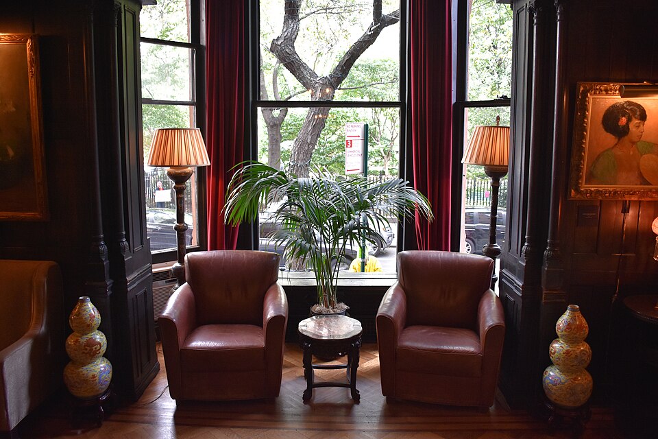 A wood paneled sitting room at the National Arts Club, leather armchairs either side of a bay window whose curtains frame the street trees and railings across the road.