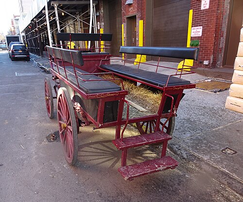 A red horse drawn wagon parked at the kerb on West 48th Street near Eleventh Avenue, hay still in the back of it.