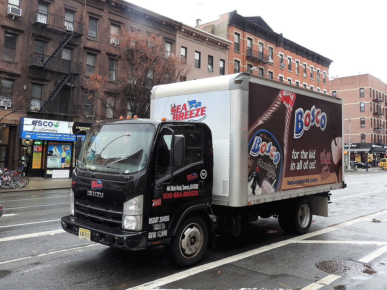 Ninth Avenue looking north from West 47th Street, a delivery truck in the roadway, brick walk-ups with fire escapes behind it and a drug store on the corner.