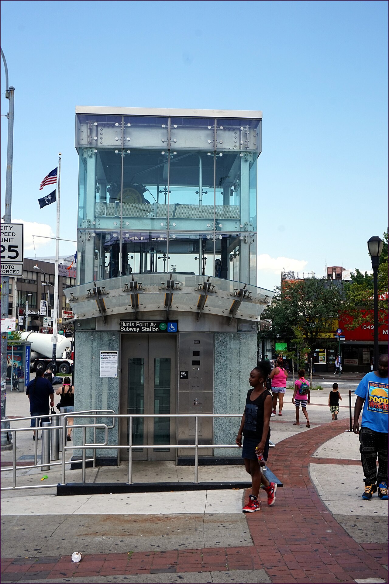 The glass elevator entrance to the Hunts Point Avenue subway station, storefronts and pedestrians on the plaza behind it.
