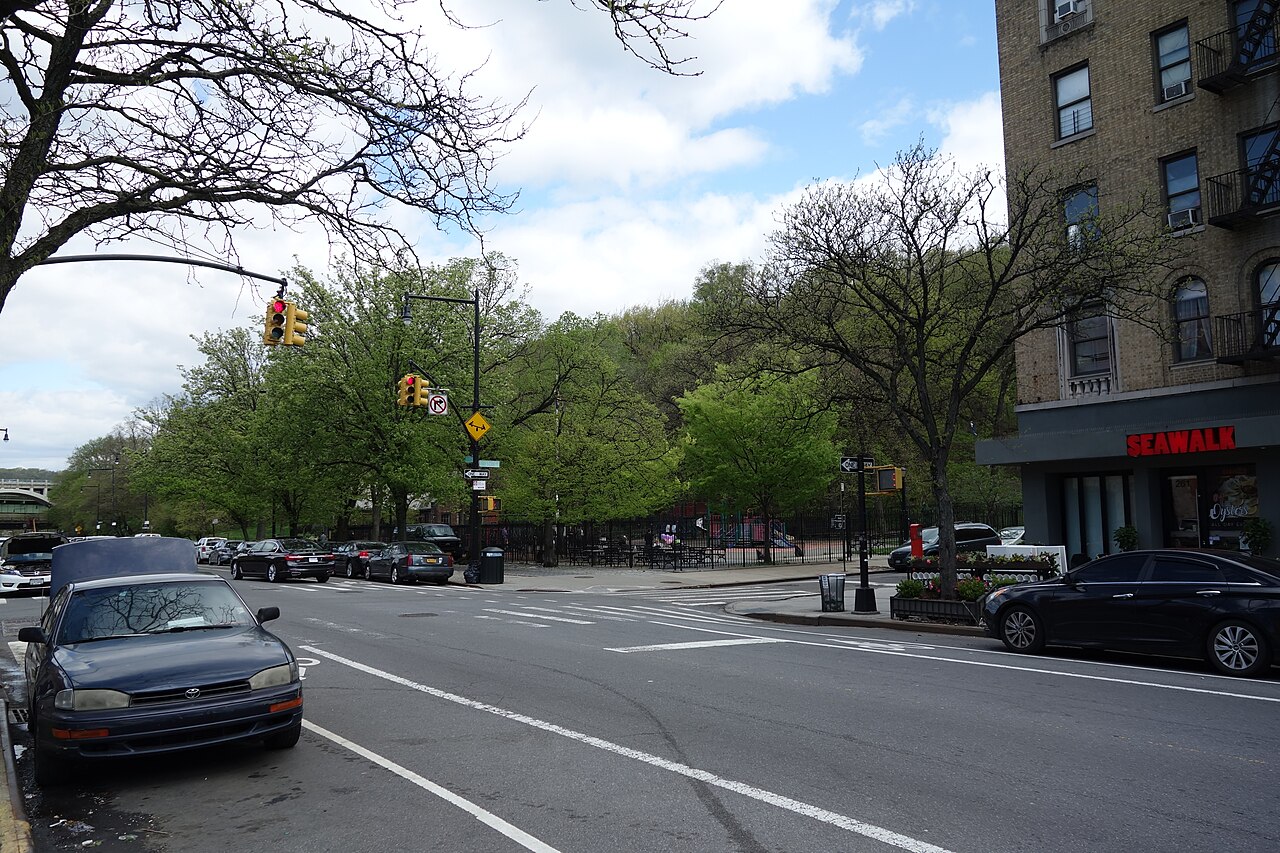 Payson Avenue at Dyckman Street, with the wooded slope of Inwood Hill Park down one side and a prewar apartment building with a storefront on the other.
