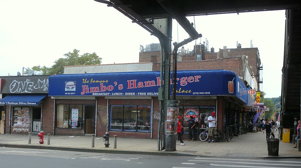 Tenth Avenue looking south from 207th Street, the elevated deck overhead and a steel column in the roadway, with a hamburger place under a long blue awning on the corner.