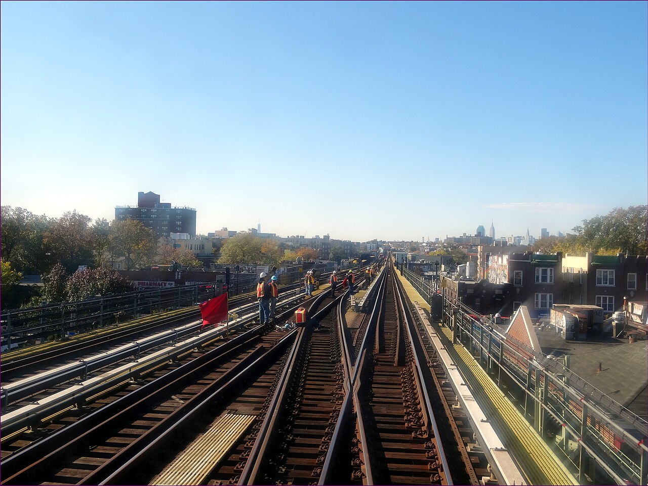 The elevated tracks of the 7 west of the 74th Street station, track workers in orange vests on the roadbed, rooftops on both sides and the Manhattan skyline in the distance.