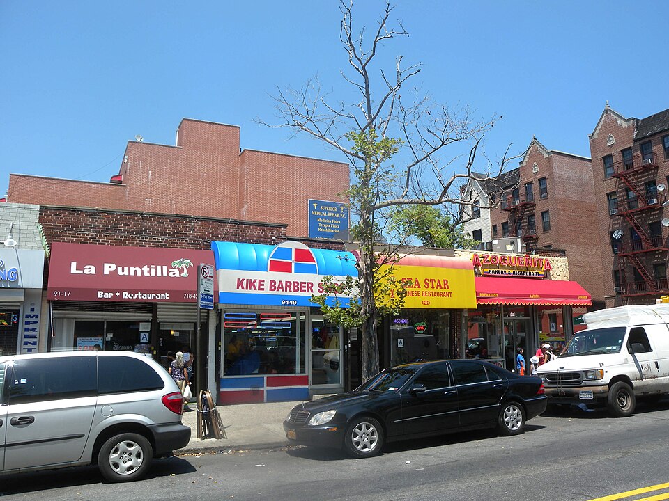 Storefronts on the north side of 37th Avenue at 91st Street, a bar, a barber shop, a Chinese restaurant and a bakery under separate awnings, brick apartment houses behind.