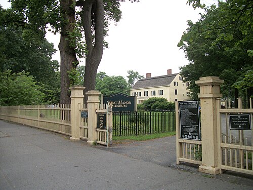 The gate into Rufus King Park, a sign reading King Manor Museum, and the wood frame house behind the fence among summer trees.