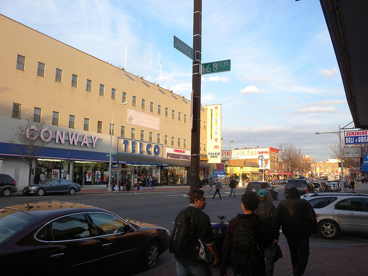 Jamaica Avenue at 168 Place looking northeast, discount stores called Conway and Telco filling the ground floors, pedestrians crossing under a green street sign in late afternoon light.