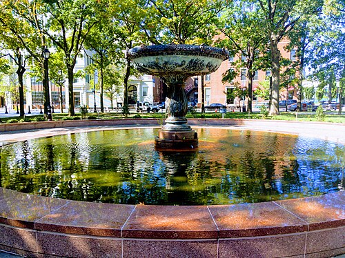 The fountain in the park at Court Square, an ornate bowl in a granite basin with trees and low brick buildings around it.