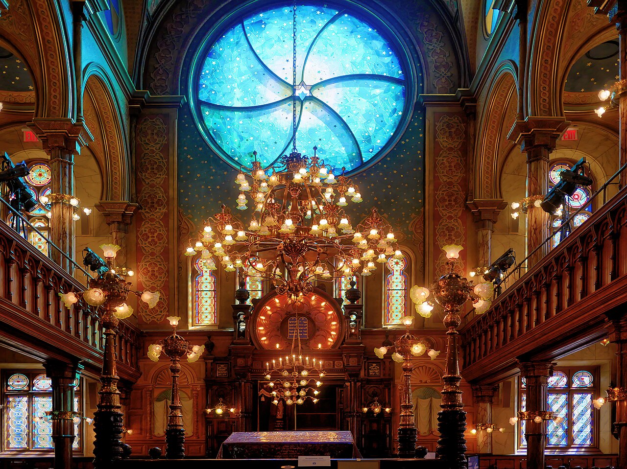 The sanctuary of the Eldridge Street Synagogue, the ark below and the blue rose window above it, with brass chandeliers hanging in front of painted arches and two levels of wooden balconies.