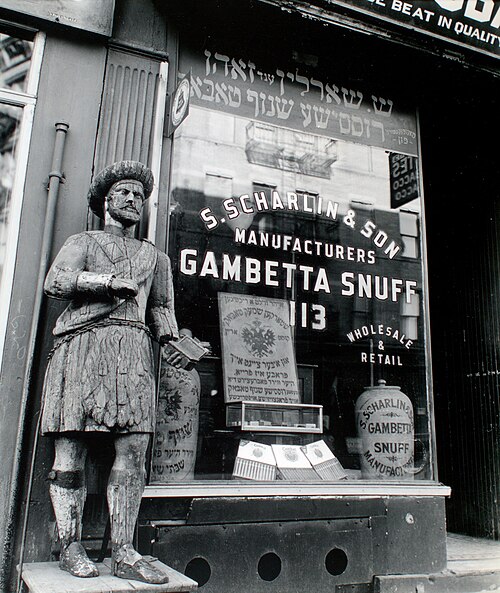 A snuff shop at 113 Division Street photographed by Berenice Abbott in 1938, a carved wooden Scotsman standing on the step and the window lettered in English and Yiddish.