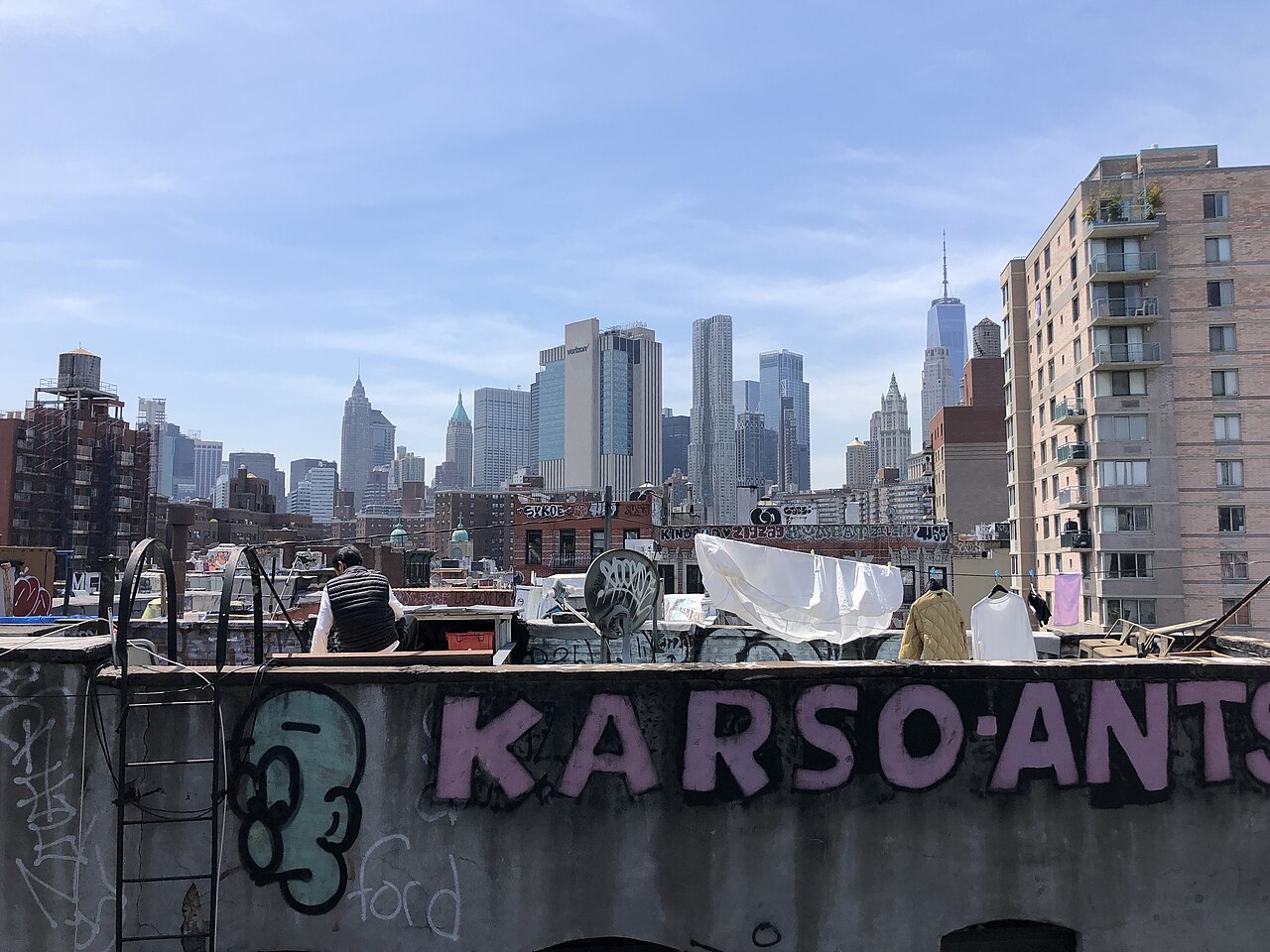 Tenement rooftops seen west from the Manhattan Bridge, washing hung on a line and a graffitied parapet in front, a man sitting out on the roof and the Financial District behind.
