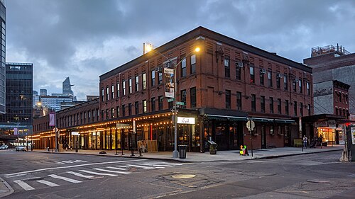 A three storey brick market building on the corner of Gansevoort and Washington Streets at dusk, string lights over an arcade of ground floor storefronts.