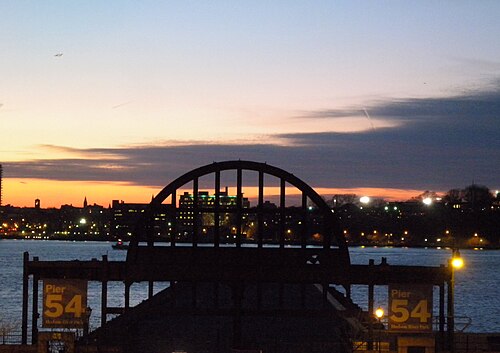 The surviving steel entrance arch of Pier 54 against the Hudson River at dusk, the New Jersey shoreline lit up behind it.